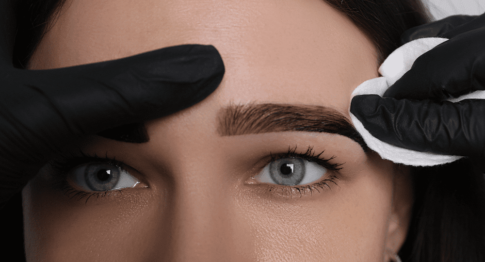 Woman getting eyebrows gently cleaned by a professional with gloves and a cotton pad.